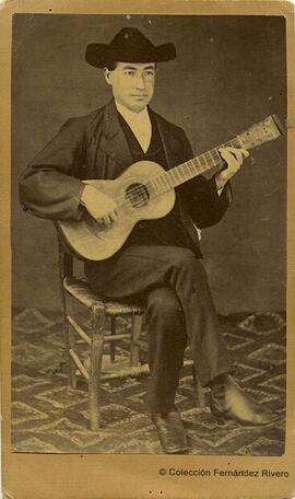 España, retrato de un guitarrista tocado con un sombrero catite y sentado en una silla de enea. Fotógrafo desconocido.