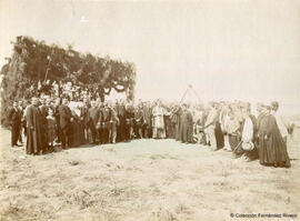 Retrato de un grupo de personas en el campo junto a un cobertizo engalanado: autoridades eclesiásticas y civiles con hermanas de la caridad y fieles. Fotógrafo desconocido.